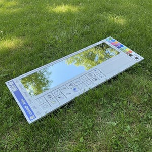 May include: A rectangular mirror designed to resemble the Windows Paint program interface. The mirror is light gray with a reflective surface, featuring a toolbar with icons and a color palette. The mirror is lying on a bed of green grass.