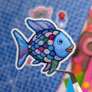 May include: A pixel art sticker of a fish with a light blue head and orange lips. The body is covered in a mosaic of multicolored scales in shades of blue, purple, pink, and green. The fins are a mix of blue and purple tones with a white border.