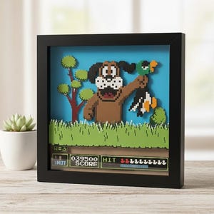 May include: A framed pixel art scene featuring a brown dog holding a green duck, reminiscent of a classic video game. The artwork includes a blue sky background, green grass, and a game interface at the bottom. The frame is black.