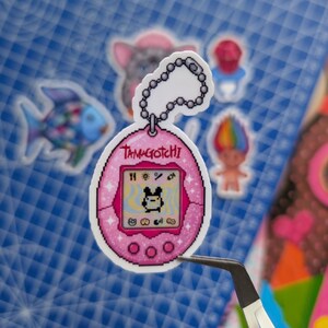 May include: A pink Tamagotchi sticker featuring a pixelated screen with a small character, a silver ball chain attachment at the top, and a glittery finish. The device has three circular buttons at the bottom and the word Tamagotchi printed above.