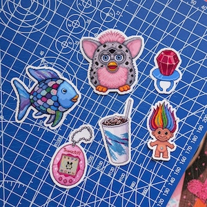 May include: A collection of five pixel art stickers featuring a gray and pink Furby, a rainbow-scaled fish, a red ring pop, a pink Tamagotchi with the text Tamagotchi on the screen, a paper cup with a straw, and a troll doll with rainbow hair.