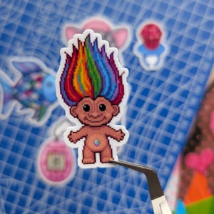 May include: A pixel art sticker of a troll doll with vibrant rainbow-colored hair and a small, iridescent gem on its belly. The figure has a tan complexion, large black eyes, and a wide smile, presented in a die-cut shape with a white border.