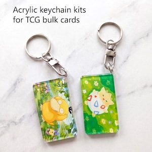 Könnte beinhalten: Zwei rechteckige Acryl-Schlüsselanhänger-Sets für TCG-Sammelkarten. Eines zeigt eine gelbe Enton-Figur in einer grünen Blumenlandschaft, das andere eine Togepi-Figur auf grünem Grund. Beide haben Schlüsselringe und Karabiner aus silbernem Metall.