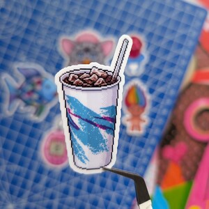 May include: A pixel art sticker of a white disposable cup filled with a dark brown iced beverage and a straw. The cup features a classic teal and purple abstract brushstroke pattern. The sticker is held by black tweezers against a blue grid mat.