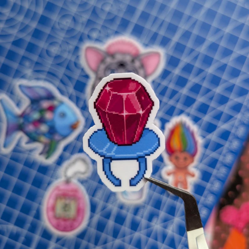 May include: A pixel art style sticker of a ring with a large, faceted, deep red gemstone set on a light blue base. The sticker is held by black metal tweezers, with other various pixel art stickers visible in the blurred background.