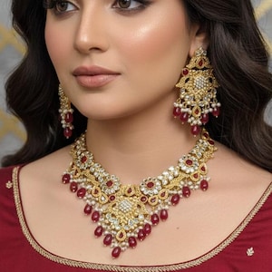 Könnte beinhalten: Ein goldfarbenes Halsketten- und Ohrring-Set mit roten und weißen Akzenten. Die Halskette hat ein zentrales Design mit roten Steinen und hängenden roten Tropfen. Die Ohrringe spiegeln das Design der Halskette wider. Die Frau trägt ein weinrotes Oberteil.