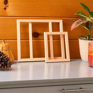 May include: Two light-colored wooden frames with a rectangular design, positioned side-by-side. The frames are empty and feature a simple, clean construction. They are displayed alongside a brown pinecone, a small orange pumpkin, a potted plant, and a lit candle.
