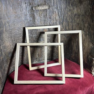 May include: Three unfinished wooden picture frames of varying sizes are arranged on a burgundy surface. The frames are rectangular with a simple, clean design, and the wood has a natural, light color. The background is a textured, dark wood.