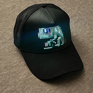 May include: A black baseball cap with a mesh back and a solid front panel featuring a glowing LED pixel art display of a dinosaur. The cap has a curved brim and a structured crown, with the digital light display emitting a bright cyan and white glow.