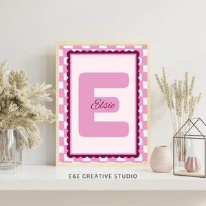 May include: A rectangular framed print featuring a large pink letter E centered on a light pink background. The design includes a dark pink scalloped border and a light pink and white checkered outer frame. The name Elsie is written in script.
