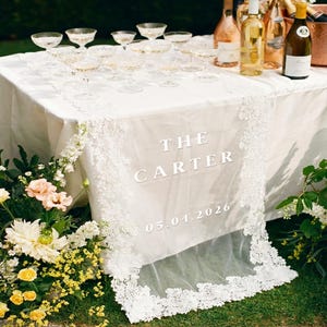 Peut inclure: Une table dressée pour une célébration, avec des coupes de champagne et des bouteilles de vin. Un chemin de table blanc avec une bordure en dentelle porte l'inscription "THE CARTER 05.04.2026". Des fleurs blanches, jaunes et roses ornent la table.