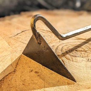 May include: Close-up of a metal gardening tool with a triangular blade and a curved handle. The tool is resting on a wooden surface. The tool has the letters "H. U.S.A." stamped on the blade.