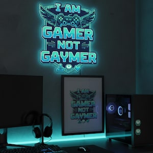 Op de afbeelding: Een blauw en turquoise neonbord met de tekst "I AM GAMER NOT GAYMER", met een controller- en vleugelontwerp. Een ingelijste print van hetzelfde ontwerp staat eronder. De scène omvat een computeropstelling met een monitor, toetsenbord en koptelefoon.