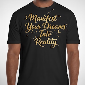 May include: Black t-shirt with gold script that reads "Manifest Your Dreams Into Reality." The design includes constellation-like star patterns and a crescent moon. The shirt is a crew neck style.