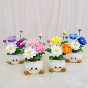 Op de afbeelding: Een verzameling handgemaakte gehaakte bloempotten, elk met een lachend gezicht en kleine bruine voetjes. De bloemen zijn in verschillende kleuren, waaronder wit, roze, geel, oranje, blauw en paars, met groene bladeren. De potten zijn wit.