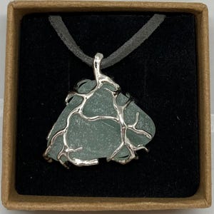 May include: A sea glass pendant necklace. The pendant is a light blue, irregularly shaped piece of sea glass, encased in a silver-colored, organic-looking wire frame. It hangs from a gray suede cord, presented in a brown box with a black interior.