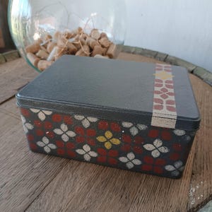 Può includere: Una scatola rettangolare in metallo grigio scuro con un motivo floreale rosso, bianco e giallo. Una fascia decorativa con un motivo floreale ripetuto in rosso e giallo adorna il coperchio. La scatola è su una superficie di legno.