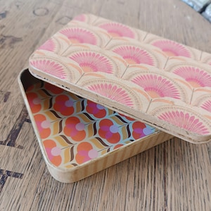 Può includere: Una scatola rettangolare aperta con coperchio. Il coperchio e l'interno presentano motivi floreali colorati in rosa, arancione e blu su sfondo beige. La scatola è appoggiata su una superficie di legno.