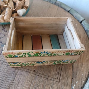 Può includere: Una piccola cassa di legno rettangolare con un motivo floreale decorativo in verde, blu e arancione. Le doghe interne della cassa sono dipinte in vari colori. La cassa è in legno chiaro.