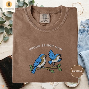 Może przedstawiać: Koszulka w kolorze espresso z wyhaftowanym wzorem dwóch niebieskich ptaków siedzących na brązowej gałęzi. Napis "PROUD SENIOR MOM" jest wyszyty białą nicią nad ptakami. Koszulka posiada metkę marki Comfort Colors przy kołnierzu.