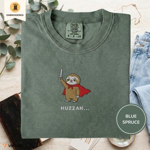 Könnte beinhalten: Blau-Fichte-T-Shirt mit einem gestickten Cartoon-Faultier, das einen roten Umhang trägt und ein Schwert hält. Der Text "Huzzah..." ist darunter gestickt. Das Shirt hat ein Comfort Colors-Etikett.