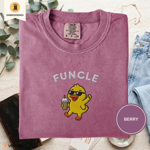 May include: A berry-colored crew neck t-shirt featuring an embroidered graphic of a yellow duck wearing black sunglasses and holding a coffee cup. The text "FUNCLE" is embroidered in white above the duck. The shirt has a Comfort Colors brand label.