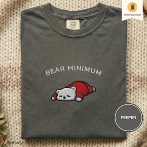 Puede incluir: Una camiseta gris oscuro con la frase bordada "BEAR MINIMUM" sobre un oso de dibujos animados con un atuendo rojo. La camiseta está doblada cuidadosamente, con una etiqueta visible en el cuello. La palabra "EMBROIDERED" está impresa en una pequeña pegatina circular.