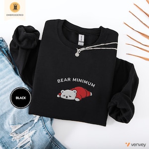 May include: A black crewneck sweatshirt featuring an embroidered graphic of a small white bear wearing a red sleeping bag, positioned below the text "BEAR MINIMUM" in white thread. The sweatshirt has long sleeves and a ribbed neckline.