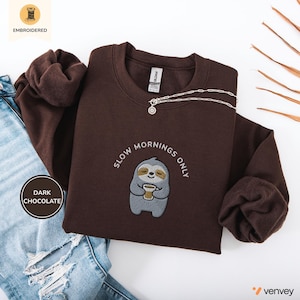 Könnte beinhalten: Ein dunkelbraunes Sweatshirt mit Rundhalsausschnitt und einer Stickerei eines grauen Faultiers, das eine Kaffeetasse hält. Der Text "SLOW MORNINGS ONLY" ist in weißem Garn bogenförmig über dem Faultier gestickt. Das Kleidungsstück hat lange Ärmel.
