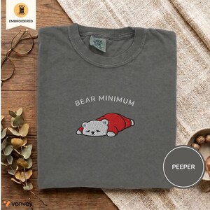 May include: Dark gray t-shirt with the embroidered phrase "BEAR MINIMUM" above a cartoon bear wearing a red outfit. The shirt has a crew neck and a relaxed fit. The word "PEEPER" is in a circle on the bottom right.
