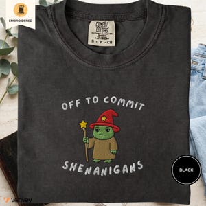 May include: A black t-shirt featuring an embroidered graphic of a green creature wearing a red wizard hat with a yellow star, holding a staff with a yellow star. The text "OFF TO COMMIT SHENANIGANS" is embroidered in white above and below the graphic.