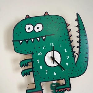 May include: A green dinosaur-shaped wall clock with a white clock face and black hands. The clock features a cartoon design with white teeth, eyes, and numbers. The clock is made of wood and has a tail.