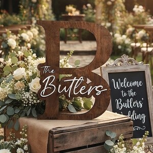 May include: A large wooden letter B with the text The Butlers in white script, placed on a wooden crate. Beside it stands a framed chalkboard sign with the text Welcome to the Butlers' wedding, surrounded by white roses and eucalyptus greenery.