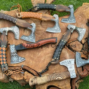 Könnte beinhalten: Eine Sammlung von Wikingeräxten mit Holzgriffen und aufwendig gestalteten Metallklingen. Die Griffe sind mit Leder umwickelt und weisen dekorative Elemente auf. Die Äxte werden auf einer Holzoberfläche mit dem Text "BLADE ZONE CREATIONS" präsentiert.