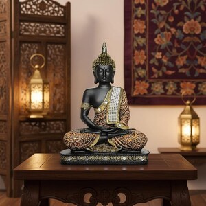 K&ouml;nnte beinhalten: Eine schwarze Buddha-Statue in sitzender Meditationshaltung. Die Statue hat goldene Akzente an Kopf, Armen und Kleidung. Die gemusterte Kleidung ist braun und goldfarben. Die Statue steht auf einem dekorativen Sockel.