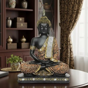 K&ouml;nnte beinhalten: Eine schwarze Buddha-Statue in sitzender Meditationshaltung. Die Statue hat goldene Akzente an Kopf, Arm und Kleidung. Das Gewand ist mit orangefarbenen und goldenen Mustern versehen. Die Statue steht auf einem schwarzen Sockel mit dekorativen Spiegelakzenten.