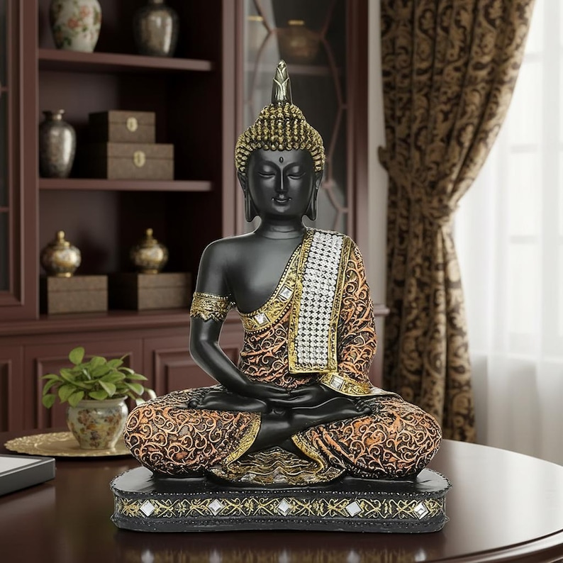 K&ouml;nnte beinhalten: Eine schwarze Buddha-Statue in sitzender Meditationshaltung. Die Statue hat goldene Akzente an Kopf, Arm und Kleidung. Das Gewand ist mit orangefarbenen und goldenen Mustern versehen. Die Statue steht auf einem schwarzen Sockel mit dekorativen Spiegelakzenten.