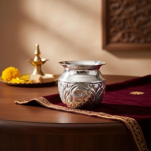 May include: A silver-colored, ornate metal pot with a wide rim and floral design. The pot sits on a dark red cloth with a gold border, on a wooden table. A brass lamp and yellow flowers are in the background.