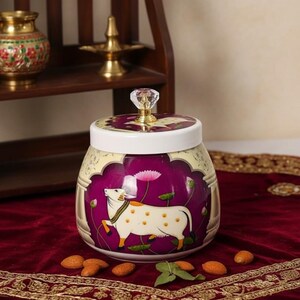 Op de afbeelding: Een decoratieve keramische pot met een witte deksel en een kristallen knop. De pot heeft een paarse achtergrond met een witte koe-illustratie en lotusbloemen. De pot staat op een rode fluwelen ondergrond.