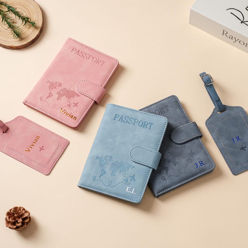 May include: Three passport holders and two luggage tags in pink, light blue, and dark blue. Each item features a world map print, the word PASSPORT, and personalized initials or names including Vivian, E.L., and J.R. with small airplane icons.