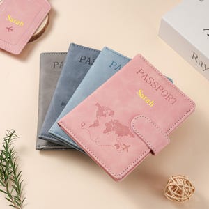 May include: A stack of three passport holders in pink, blue, and gray. Each holder features the word PASSPORT, a world map graphic, and a small airplane icon. The pink holder is personalized with the name Sarah in gold lettering.