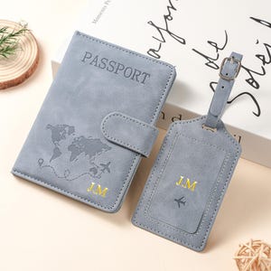 May include: A grey passport holder and matching luggage tag set. The passport holder features the word PASSPORT, a world map graphic, and the gold embroidered initials J.M. The luggage tag includes the gold initials J.M and a small airplane icon.