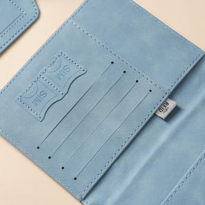 May include: A light blue passport holder featuring interior slots for cards and two dedicated SIM card pockets. The interior includes a small fabric tag with the text RFID printed on it, indicating the presence of radio-frequency identification protection.