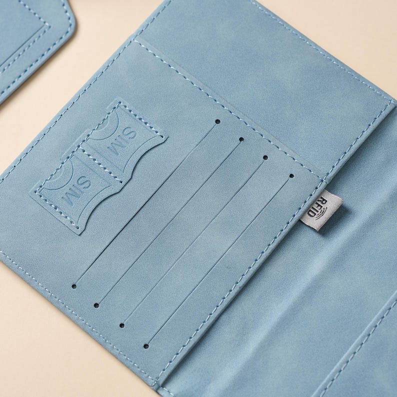 May include: A light blue passport holder featuring interior slots for cards and two dedicated SIM card pockets. The interior includes a small fabric tag with the text RFID printed on it, indicating the presence of radio-frequency identification protection.