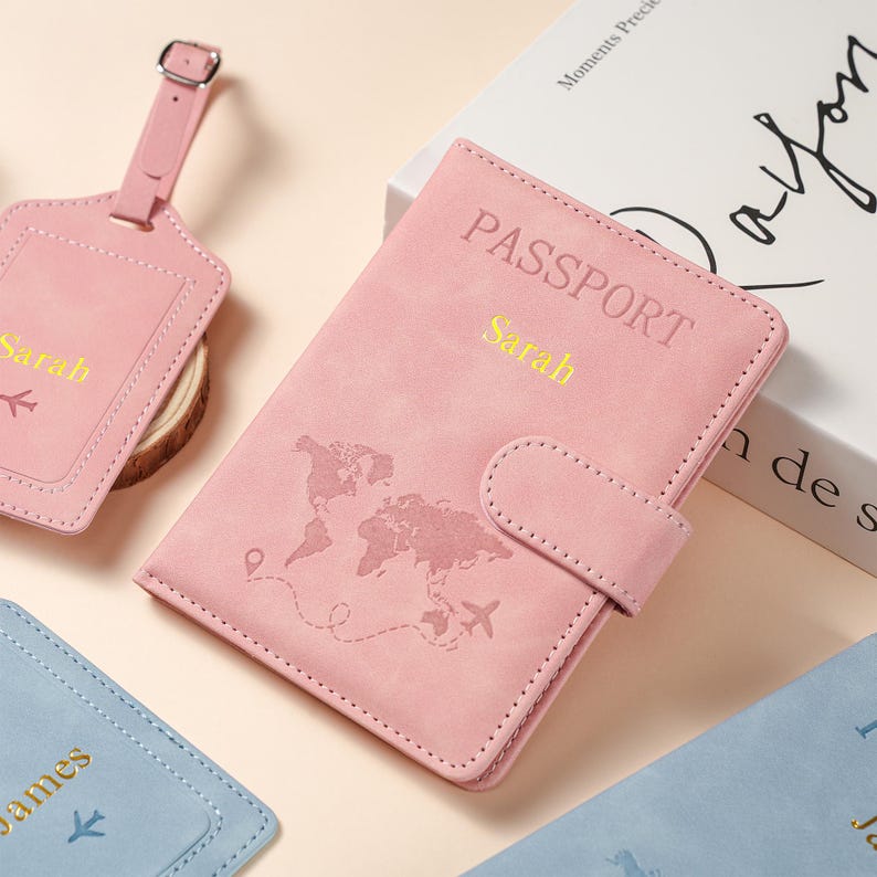 May include: A pink passport holder with a world map graphic, a small airplane icon, and the name Sarah in gold lettering. A matching pink luggage tag with the name Sarah and an airplane icon is included, alongside a blue luggage tag with the name James.