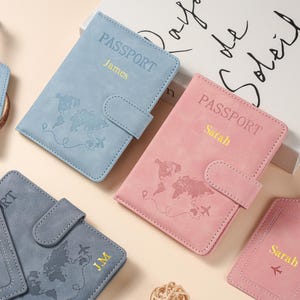 May include: A collection of passport holders in light blue, pink, and dark gray, each featuring a world map print, a snap closure, and personalized names or initials in gold lettering. The covers are textured and include a small airplane icon.