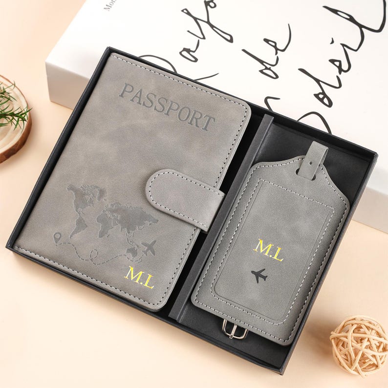 May include: A gray passport holder and luggage tag set in a black gift box. The passport holder features a world map, the word PASSPORT, and the initials M.L. in gold. The luggage tag includes the initials M.L. and a small airplane icon in gold.