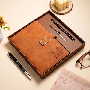 May include: A tan notebook with a gold-toned metal clasp and the name Matthew engraved on the cover, presented in a brown gift box with two pens, one of which is a black and silver ballpoint pen resting outside the box on a light surface.