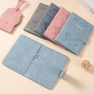 May include: A collection of faux leather passport holders in gray, blue, and pink, featuring embossed world map designs and personalized names. One light blue holder is open, revealing card slots and SIM card pockets, alongside a matching luggage tag.