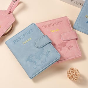 May include: Two rectangular passport holders, one light blue and one pink, featuring a snap closure, a world map embossed on the front, and the words PASSPORT with a personalized name in gold lettering. A matching luggage tag is visible.